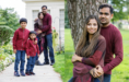 A family of four poses outdoors. On the left, parents and two boys in red sweaters stand on a garden path. On the right, the parents stand close together by a tree, smiling at the camera.