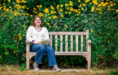 A person wearing a white sweater, blue jeans, and sneakers sits on a wooden bench holding a bouquet of wildflowers during a summer senior pictures session, with tall yellow flowers and green foliage in the background.