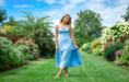 A woman in a light blue dress stands barefoot on a green lawn, smiling and holding her dress during a summer senior pictures session. She is surrounded by colorful flowers and lush greenery under a partly cloudy sky.