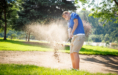 A golfer in a blue shirt hits a shot from a sand bunker, sending sand into the air on a sunny day—perfect for dynamic golf senior pictures.
