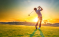 A golfer swings a club on a green golf course at sunset, with the sun low on the horizon casting a warm golden light and long shadows on the grass.