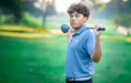 A young person stands on a golf course holding a golf club across their shoulders, wearing a light blue polo shirt and looking at the camera. The background is grassy and blurred with sunlight filtering through trees.