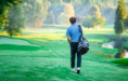 A high school senior wearing a blue shirt and black pants walks alone across a lush, green golf course carrying a golf bag over their shoulder, surrounded by trees and early morning light—perfect for a golf senior pictures session.