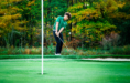 A high school senior guy in a green shirt and black pants prepares to chip a golf ball onto the green during a golf senior pictures session, with a white flag in the foreground and trees with autumn foliage in the background.