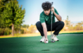 A young person in a green shirt and black pants squats on a golf course, holding a putter and positioning a golf ball on the green, with trees and blue sky in the background.