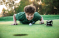 A young man in a green polo shirt lies on a golf green, face close to the ground, lining up a putt with a golf ball and putter as he smiles intensely toward the hole in the foreground.