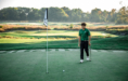 A young golfer in a green shirt and black pants stands on a golf green, holding a putter and looking at a golf ball near the hole, with trees and fairways in the background.