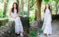 A high school senior girl in a white floral dress poses outdoors beside a stone wall and walks along a sunlit forest path, surrounded by green trees and foliage—perfect inspiration for Grosse Pointe senior pictures.