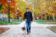 A young man in a black shirt and jeans walks a brown and white dog on a leash along a tree-lined path, surrounded by colorful autumn leaves—an ideal scene for a fall senior pictures session.
