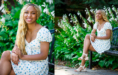 A woman with long blonde braids, wearing a white floral dress and sandals, sits and smiles on a bench in a garden with green foliage and white flowers—the perfect setting for a summer senior pictures session.