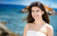 A young woman with long brown hair, wearing a white strapless top, smiles brightly during her summer senior pictures session outdoors near a blue ocean with rocky outcrops in the background on a sunny day.