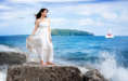 A high school senior girl in a white dress stands on the rocky shores of Mackinac Island, where waves splash around her. A seagull flies nearby, and a lighthouse is visible on the horizon under the expansive blue sky—an ideal spot for senior pictures.