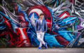 A young man in a blue blazer, white shirt, and white pants leans against a colorful graffiti-covered brick wall, looking off to the side with hands in his pockets.