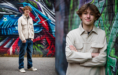 A teenage boy with brown hair, wearing a beige jacket, blue jeans, and sneakers, stands and poses in front of a colorful graffiti wall, smiling confidently at the camera.