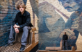 A young man with light brown hair, wearing a black jacket and gray jeans, sits and stands on a modern wooden bench against a brick wall mural depicting a large, detailed face and arm.