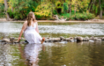 During a summer senior pictures session, a woman in a white dress sits on rocks in a shallow river, smiling and splashing water with her feet. Trees with green and yellow leaves are visible in the background.