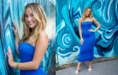 A young woman with long blonde hair wears a strapless blue dress and white sandals, smiling while posing in front of a vibrant blue and turquoise graffiti mural.