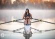 A high school senior in athletic gear is rowing a single scull on calm water during a misty morning, reminiscent of serene senior pictures. She is holding oars with focused determination, set against a backdrop of soft, mist-covered trees.