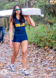 A high school athlete in a navy uniform with SALINE printed on it carries a rowing boat on her shoulder. She's part of the rowing crew, walking on a leaf-covered path in a wooded area, wearing sunglasses and smiling, capturing the perfect senior picture.