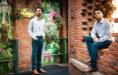 A high school senior guy an in a checked shirt and jeans poses for senior photographers in a brick-walled room adorned with lush green plants, chandeliers, and a vibrant, eclectic decor.