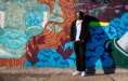 A person in a black jacket and pants stands against a colorful graffiti-covered wall, with sunlight casting their shadow beside them. The mural features abstract shapes, lips, clouds, and flowing lines.