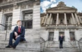 A person in a suit with a red tie sits on stone steps in front of a large, ornate building in one image. In another image, the person stands on the steps, looking at the camera, with the building's grand façade in the background.
