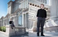A person with red hair in a black shirt and gray pants poses outside a large, ornate building with tall windows. One photo shows them sitting on a stone ledge, and the other shows them standing under a row of lights.