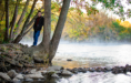 A young person stands on rocks by the base of a tree next to a calm river, surrounded by autumn foliage during their fall senior pictures session. The water reflects the colorful leaves as morning mist rises in the background.