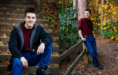 A young man with short brown hair wearing a red plaid shirt, jeans, and a black jacket poses for his fall senior pictures session, sitting on leafy outdoor steps in one photo and leaning on a wooden fence in a wooded area in the other.