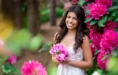 A high school senior girl in a floral dress stands outdoors, smiling and holding pink flowers—surrounded by vibrant pink blossoms and greenery. This scene captures the perfect vibe for senior pictures with flowers, set against a beautifully blurred natural background.