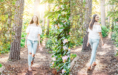 A young woman in a white off-shoulder top and ripped jeans walks along a sunlit forest path during her spring senior pictures session, smiling. One image shows her facing forward, the other captures her walking away through greenery and flowers.