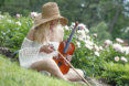 A woman in a white, off-the-shoulder dress and straw hat sits on the grass in a garden, surrounded by blooming flowers, tuning her violin during a dreamy Spring Senior Pictures session.