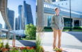 A young man in a light green shirt and tan pants stands outdoors near flower beds, with tall modern skyscrapers and a covered walkway in the background on a sunny day during his spring senior pictures session.
