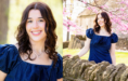 A young woman with wavy brown hair, wearing a blue dress, smiles outdoors during her Spring Senior Pictures session. In one image, she is close-up; in the other, she stands by a stone wall with blooming trees and a rustic building in the background.