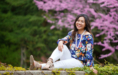 A woman sits outdoors on a stone ledge, smiling during her spring senior pictures session. She wears a floral off-shoulder blouse, white pants, sandals, and leis. Pink blossoms and green trees fill the background.