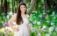 A young woman in a white dress stands smiling in a sunlit garden filled with purple and white flowers, green trees in the background—capturing the joy of her Spring Senior Pictures session.