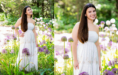 A young woman with long brown hair wearing a white dress stands and smiles among tall purple and white flowers in a sunlit garden, capturing the beauty of her Spring Senior Pictures session amid lush green foliage.
