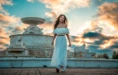 A high school senior girl in a white off-shoulder top and long skirt walks and smiles in front of a large ornate fountain at sunset, with dramatic clouds and warm sunlight in the sky behind her.