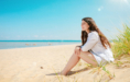 Young woman with long brown hair sits on sandy beach near grass, smiling and looking at the sea under a bright blue sky—perfect for a summer senior pictures session.