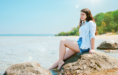A young woman with long brown hair sits on a large rock by the beach during her summer senior pictures session, barefoot in a light blue shirt and denim shorts. Calm water and a tree-lined shoreline stretch beneath the clear, sunny sky.
