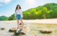 A young woman stands barefoot on rocks in shallow water at a sunny beach during her summer senior pictures session, wearing a white tank top, denim shorts, and an open shirt. Green trees and hills appear in the background under a bright sky.