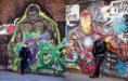 A man in a black varsity jacket and boots stands against two colorful graffiti walls—one featuring the Hulk and the other Iron Man—posing with a relaxed expression.